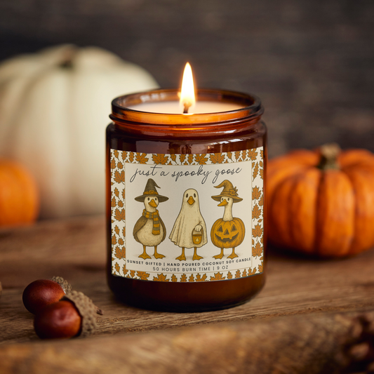 Candle with a decorative label featuring cartoon ghosts and pumpkins, surrounded by pumpkins and chestnuts on a wooden surface.